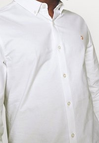 Bílá košile s knoflíky a malým zlatým vyšívaným emblémem na levé kapse, kterou nosí osoba s tmavou pokožkou.