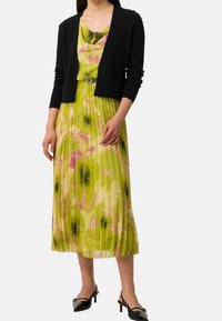 Černý kardigan přes květinové šaty v zelených, růžových a žlutých odstínech. Šaty mají záhyby, přiléhavý pas a jemný materiál. Černé boty.