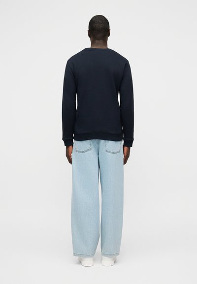 Άνδρας που φαίνεται από πίσω, φοράει μαύρο πουκάμισο με μακριά μανίκια, ανοιχτό μπλε τζιν παντελόνι με φαρδιά πόδια και λευκά αθλητικά, σε απλό φόντο.