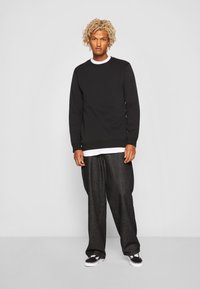 Schwarzer Sweatshirt mit Rundhalsausschnitt, strukturiertem Stoff, über einem weißen Hemd getragen, kombiniert mit weiten schwarzen Hosen und schwarzen Sneakers.