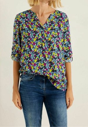Femme portant un chemisier à fleurs à manches longues avec des poignets retroussés et un jean bleu, debout devant un fond clair uni.