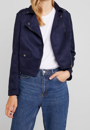 Nainen, joka käyttää tummansinistä mokkanahkaista moottoripyöräntakkia valkoisen t-paidan päällä ja korkeavyötäröisiä sinisiä farkkuja, toinen käsi taskussa, harmaalla taustalla.