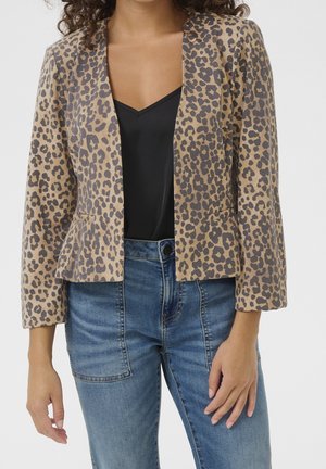 Kvinde iført en blazer med leopardprint, sort v-hals top og blå jeans, stående med armene afslappet ned langs siderne mod en hvid baggrund.