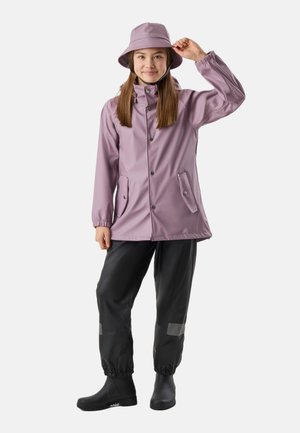 Giovane con giacca da pioggia malva e cappello, pantaloni impermeabili neri con toppe riflettenti e stivali da pioggia neri, in piedi su sfondo bianco.