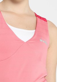 Ljust rosa sportstopp med V-ringning, texturerat tyg och kontrasterande röda remmar. Inkluderar ett vitt innerlager och subtil brandingdetalj.