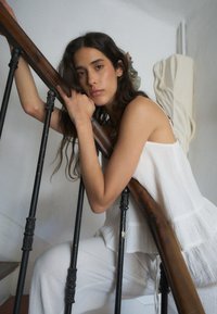 Model in einem weißen strukturierten Oberteil, der auf einem Holzhandlauf ruht. Schwarzes Metallgeländer mit dekorativen Akzenten. Sanfter, neutraler Hintergrund.