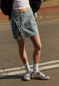 Denim wikkelrok in lichtblauw met zichtbare stiksels en knoopaccenten, gecombineerd met glanzende zilveren sneakers en witte crew-sokken.