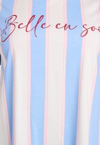 Rinnatud tunika, millel on hele sinine ja roosa vertikaalne triip valgel taustal. Tekst "Belle en soi" burgundi punase käekirjaga ees.