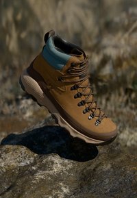 Bota de caminhada castanha com um detalhe azul, apresentando um design de tornozelo alto, couro texturizado e um sistema de atacadores durável. Projetada para uso ao ar livre.