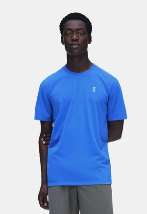 Jeune homme à la peau foncée portant un t-shirt bleu vif à manches courtes et un pantalon gris, debout devant un fond blanc uni.