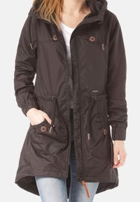 Svart huvjacka med prickigt mönster, dragsko i midjan, två framfickor och dekorativa bruna knappar. Tillverkad av ett lätt material.