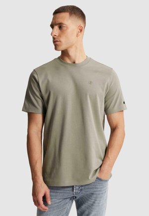 Man draagt een licht olijfgroen T-shirt met korte mouwen en een klein logo op de borst en lichtblauwe spijkerbroek, hand in de zak, kijkt naar links.