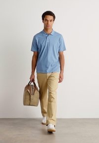 Cămașă polo albastru deschis, pantaloni bej, teniși albi și o geantă bej, mergând pe un podea netedă, pe un fundal simplu.