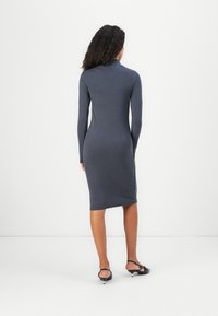 Ženska s kodrasto pričesko, oblečena v priljubljeno, dolgonogo, sivko obleko do kolen in črne čevlje z nizko peto, stoji obrnjen proti beli steni.