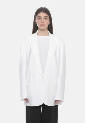 Donna con i capelli lunghi che indossa un blazer bianco oversize, una camicia bianca e pantaloni neri, in piedi davanti a uno sfondo chiaro e uniforme.