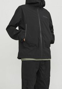 Chaqueta negra impermeable con capucha, que presenta un cierre frontal, bolsillos laterales y un corte relajado. Lleva puesta sobre una camiseta verde oscuro y pantalones a juego.