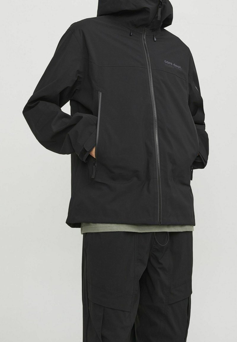 Chaqueta negra impermeable con capucha, que presenta un cierre frontal, bolsillos laterales y un corte relajado. Lleva puesta sobre una camiseta verde oscuro y pantalones a juego.