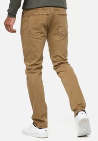 Pantalones de mezclilla de color marrón claro con un diseño de pierna recta, que cuentan con dos bolsillos traseros y una textura sutil. Llevados con zapatillas blancas.