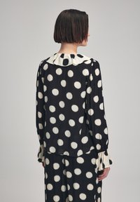 Blusa nera con grandi pois bianchi, colletto plissettato, maniche lunghe con soffici polsini e una texture di tessuto morbido e fluido.