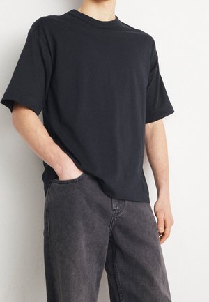 Person wearing a plain black short-sleeve t-shirt and dark gray jeans, standing with one hand in pocket against a light background.