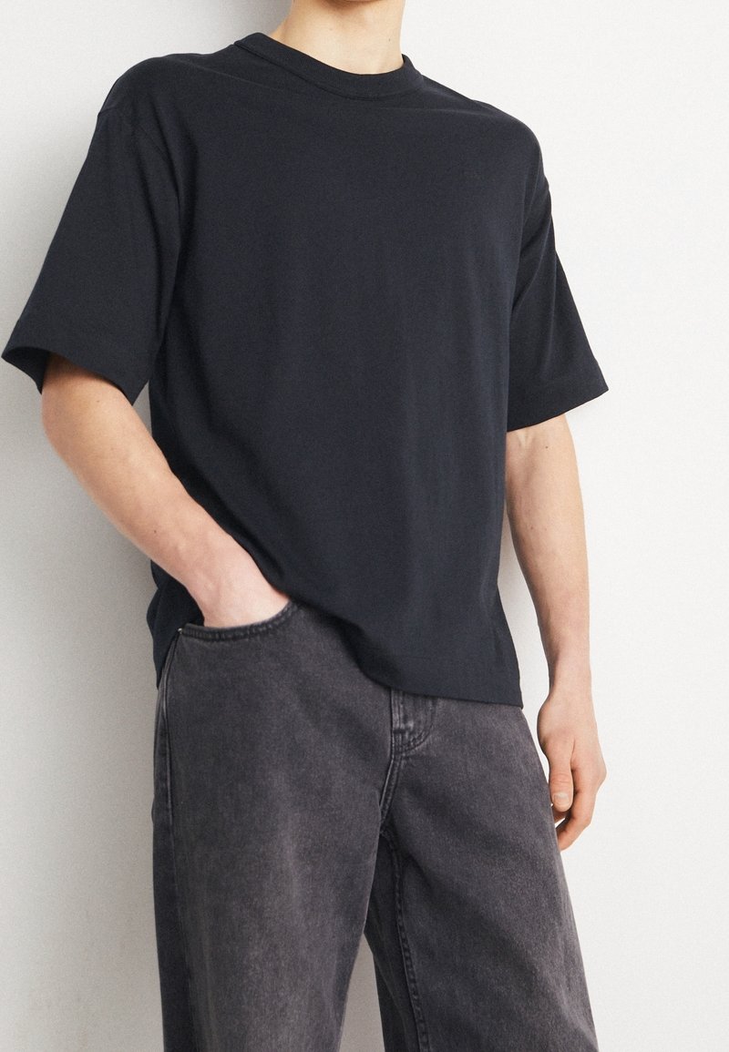 Personne portant un t-shirt noir uni à manches courtes et un jean gris foncé, debout avec une main dans la poche devant un fond clair.