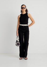 Frau trägt ein schwarzes ärmelloses Oberteil und Hose, hält eine schwarz-weiß gemusterte Handtasche, trägt eine Sonnenbrille und Sandalen.