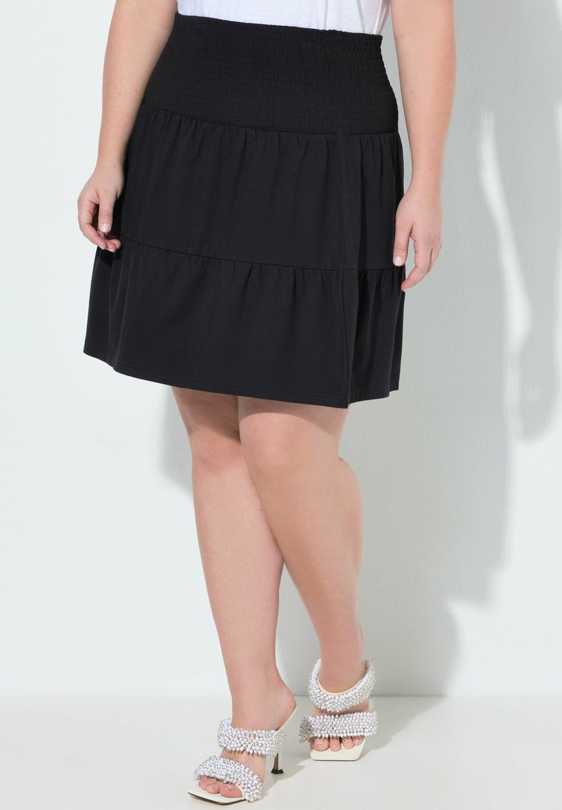 Mujer con una falda negra de volantes y cintura fruncida, y sandalias blancas de tacón alto adornadas con detalles texturizados.