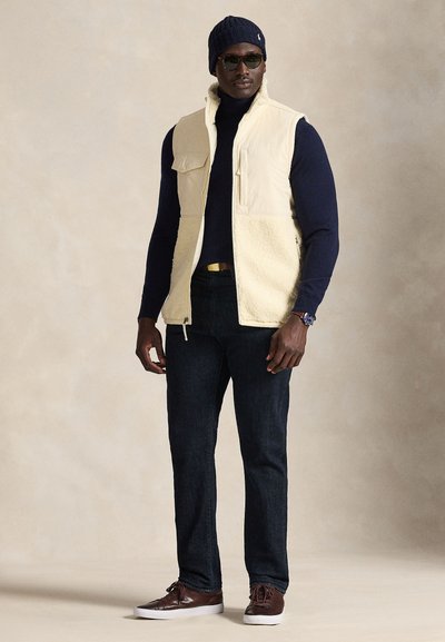 Beige herrväst med fleecefoder över en marinblå tröja, mörka jeans och bruna sneakers. Accessoarer inkluderar en marinblå mössa och solglasögon.