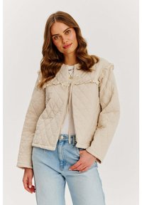 Beżowa pikowana kurtka z falbaniastym kołnierzem, z zapięciem na zamek i długimi rękawami, zestawiona z białą bluzką i jasnoniebieskimi dżinsami. Gładka tekstura.