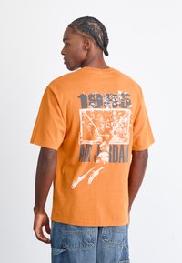 Orange bomulds T-shirt med rygdesign, der viser en basketballkurv, spillerens silhuet, "1985" og "AIR JORDAN" i sort og hvid.