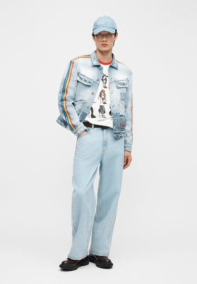 Νεαρός άνδρας που φοράει ανοιχτό μπλε τζιν μπουφάν με ρίγες ουράνιου τόξου, μπλουζάκι με σχέδιο, φαρδιά τζιν, σκούρα παπούτσια και ανοιχτό μπλε καπέλο με το κείμενο "HOT".