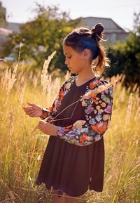 Black dress with floral patterned sleeves, braided hair, and a girl in a field of golden grass, holding a stem. Sunlight illuminates the scene.