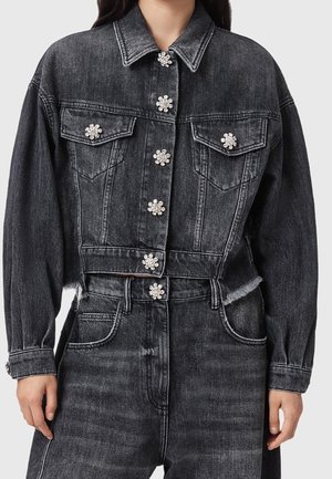Kobieta nosząca czarną, wypraną dżinsową kurtkę i spodnie z kwiatowymi guzikami z kryształków i surowymi krawędziami.