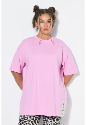 Kobieta z warkoczami, ubrana w oversize różowy t-shirt i czarno-białe spodnie w panterkę, stojąca na białym tle.