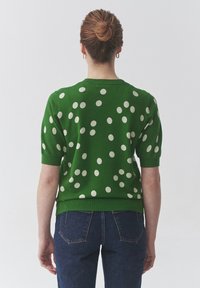 Zelený pletený svetr s krátkými rukávy, zdobený bílými puntíky a kulatým výstřihem. Kombinován s modrými džínami pro kontrast.