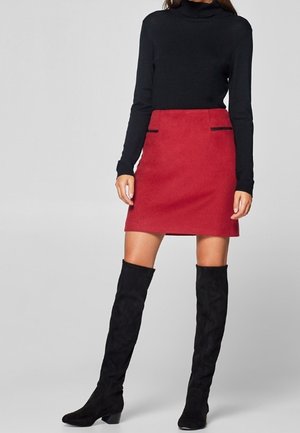 Kvinde iført en sort langærmet rullekravstrøje, rød mini nederdel med sorte lomme detaljer og sorte knæhøje støvler foran en neutral baggrund.