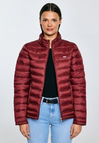 Žena v bordové prošívané bundě přes černý top a světle modré džíny, stojící před jednobarevným světlým pozadím.