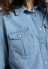 Camicia di jeans azzurro chiaro con tasche a scatto e colletto, indossata da una persona con capelli castani ondulati.