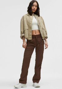Beige cropped jack met capuchon, twee voorzakken, gecombineerd met bruine elastische broek met taille en witte sneakers. Zachte stofstructuur.