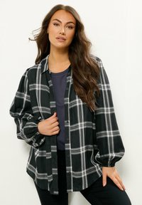 Camicia a scacchi nera e bianca con bottoni, maniche lunghe e polsini elastici, indossata sopra un top scuro; presenta una vestibilità comoda e un tessuto morbido.