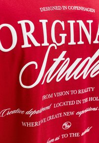 Punane puuvillane T-särk, millel on silmapaistvad valged tekstid: "Originaal Stuudio" ja lisafraasid loovuse ja asukoha kohta, erinevate fontide stiilidega.
