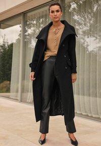 Palton lunga din lână neagră, cu guler larg, design dublu butonat, asortat cu un top tricotat bej și pantaloni din piele sintetică negri. Tocuri negre ascuțite.