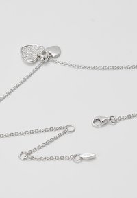 Collier en chaîne argentée avec deux pendentifs en forme de cœur, l'un incrusté de petits cristaux, avec une fermeture à pince homard sur fond blanc.