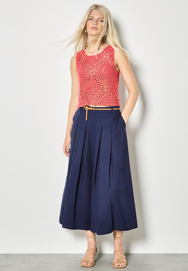 Femme portant un haut sans manches en crochet corail, une jupe midi plissée bleu marine avec une ceinture beige, et des sandales à lanières beige, debout contre un mur blanc.