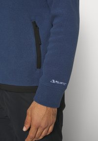 Lähikuva henkilöstä, joka käyttää sinistä Polartec-fleece-takkia, jossa on musta vetoketjullinen tasku, sekä mustia housuja.