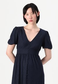 Marinblå broderad klänning med puffärmar, V-ringning och framknappar. Har texturerade blommönster och en figurnära midja.
