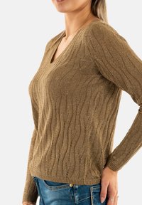 Maglione marrone, con texture, a V e con motivi a onda, realizzato in un materiale lucido. Maniche lunghe e dettagli a coste nel dorso. Indossato con jeans blu.