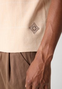 Beige strikbluse med geometrisk mønster og brun broderet blomsterdetalje på siden, kombineret med brune plisserede bukser.
