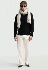 Moški v črnem puloverju, velikem beigem šalu, belih hlačah, črnih športnih čevljih in sončnih očalih, stoji pred svetlo sivo ozadjem.