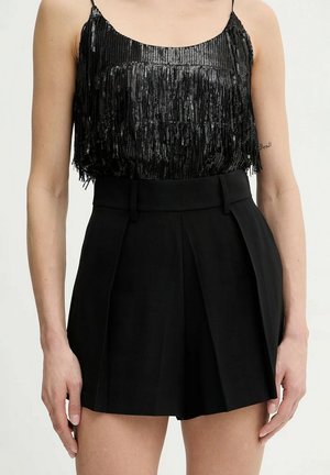 Donna che indossa un top senza maniche nero con paillettes e pantaloncini neri plissettati a vita alta, in piedi contro uno sfondo chiaro e uniforme.